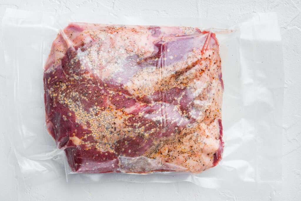 fresh-uncooked-raw-meat-beef-brisket-set-with-ingr-2026-03-26-04-34-18-utc Raw steak with pepper and herbs in a vacuum-sealed bag on a white surface.