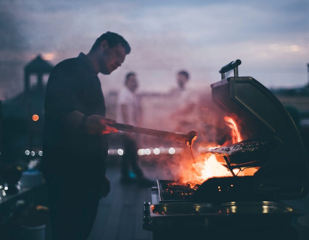 man-by-the-bbq-with-pork-chops-2026-03-10-02-50-22-utc Man tending a glowing charcoal grill on a deck at dusk, with others blurred in the background.