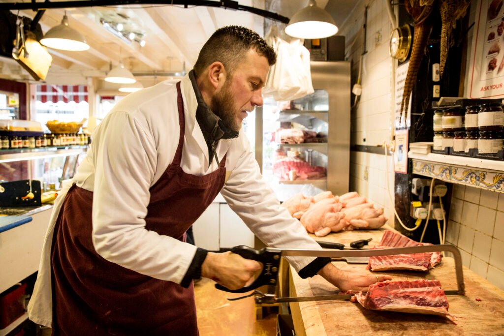 Man wearing apron standing at wooden butchers block butchering piece of meat using saw Butcher in a white coat and burgundy apron slices a ribeye with a handsaw in a meat counter.