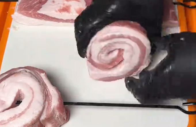 Screenshot 2026-05-29 104917 Gloved hands roll a spiral of raw pork on a white cutting board, ready for prep.