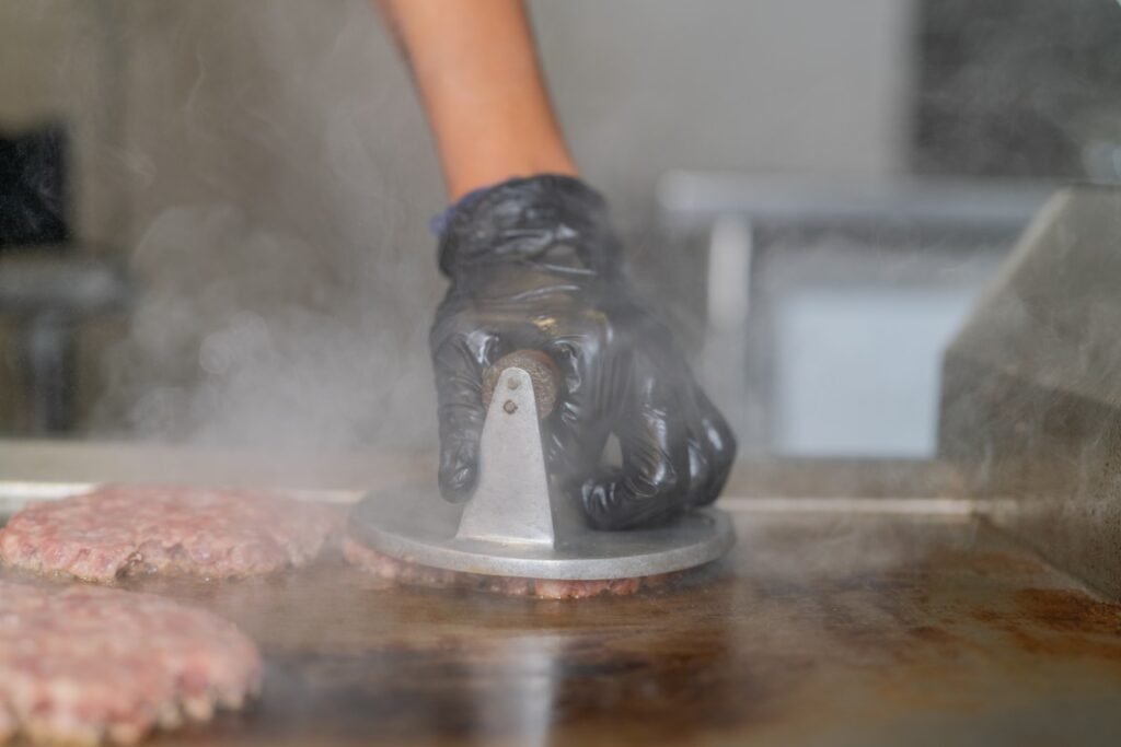 Hand in a black glove pressing a burger patty onto a hot griddle with a metal patty press, steam rising.