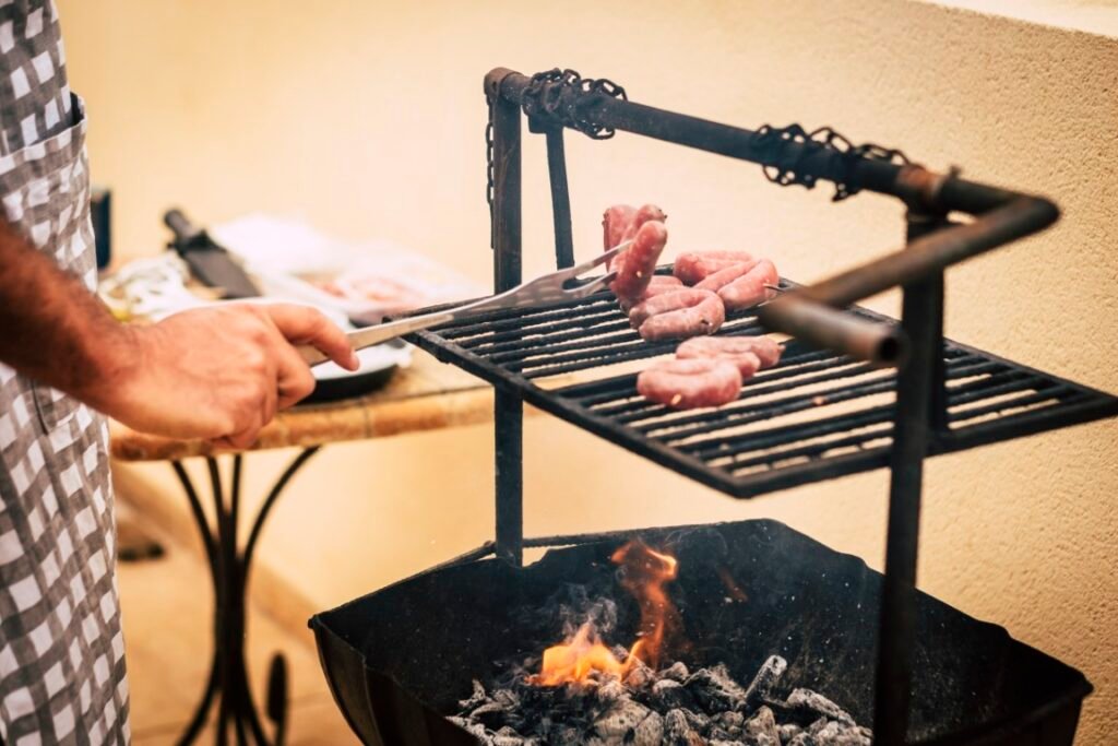 Close up of man cooking on bbq grill outdoor Home celebration a Person in a checkered shirt uses tongs to turn raw sausages on a charcoal grill over glowing coals in a small outdoor setup.