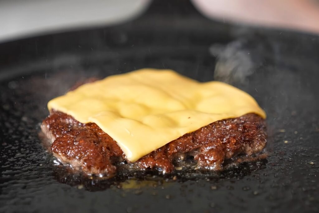 Close-up of a beef patty with melted cheese on a hot, flat-griddle surface.