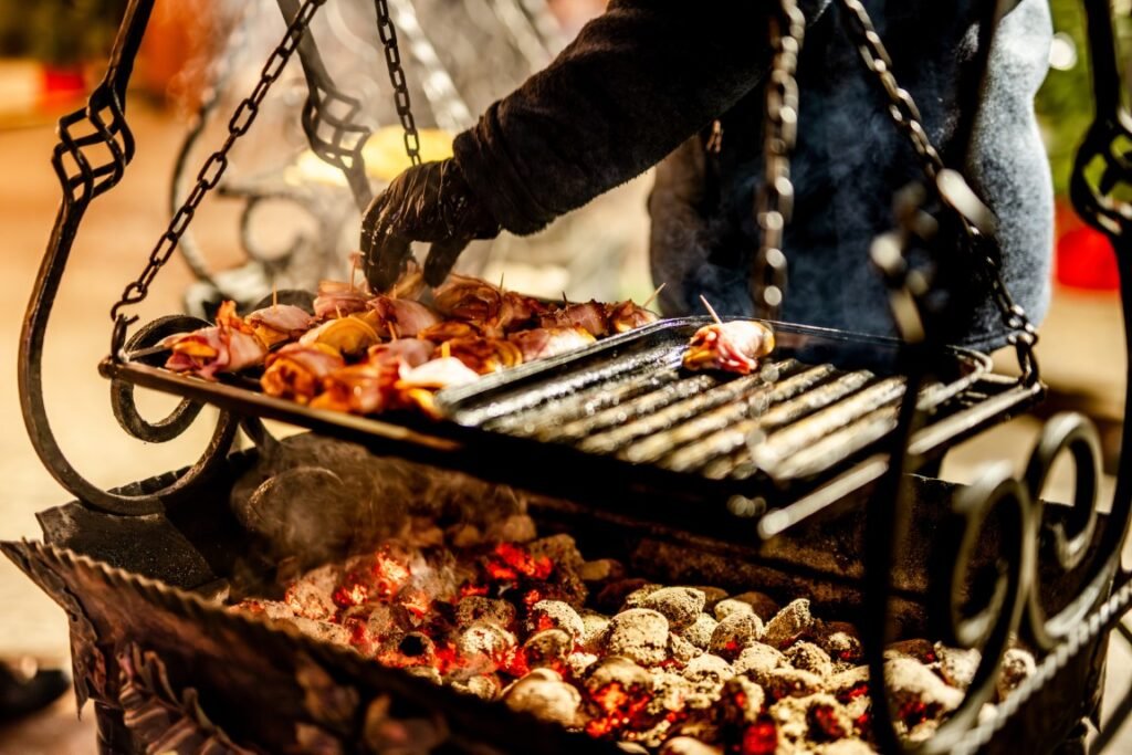 Woman Grilling Meat Snacks Person wearing black gloves grilling skewered meat over glowing charcoal in an ornate outdoor grill.