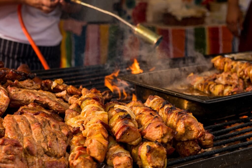 preparation BBQ Skewers of grilled meat on a sizzling open barbecue with visible flames and a colorful stall backdrop behind.