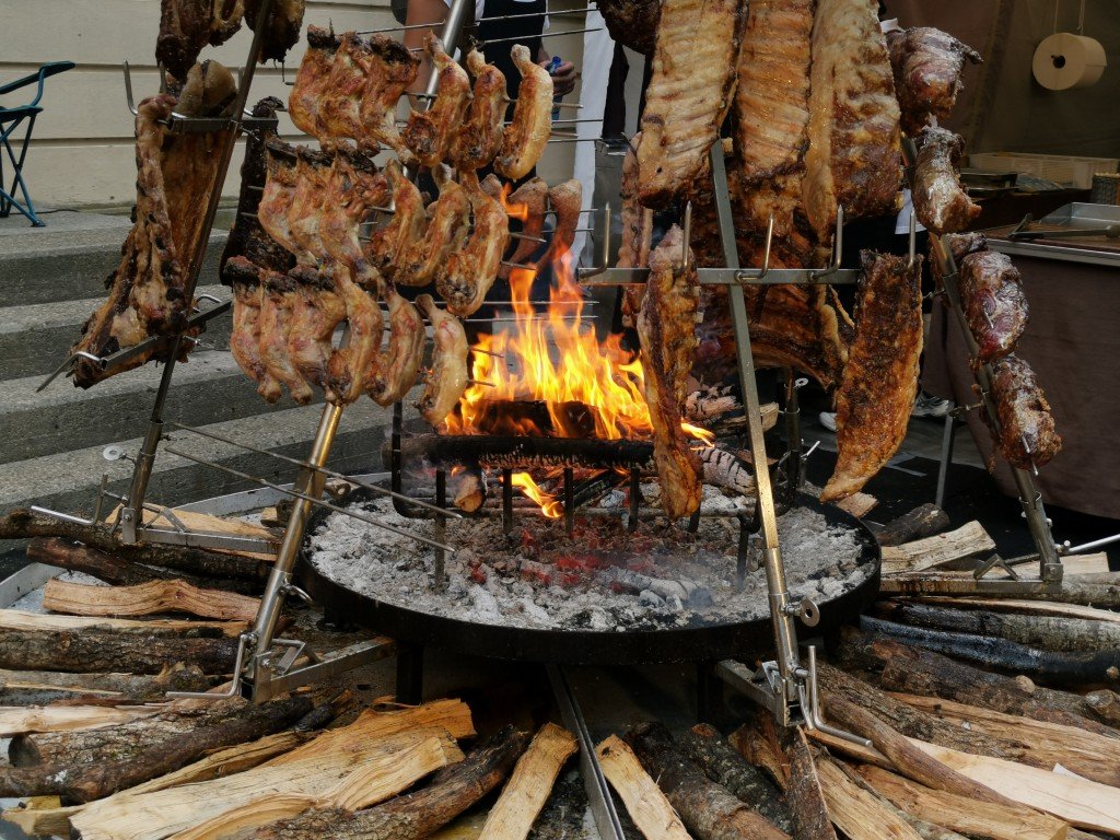 grilled-meat-preparation-over-fire-in-outdoor-sett-2026-03-16-00-38-22-utc Rotisserie-style barbecue with multiple racks of meat cooking over an open flame, surrounded by logs and a bed of hot coals.
