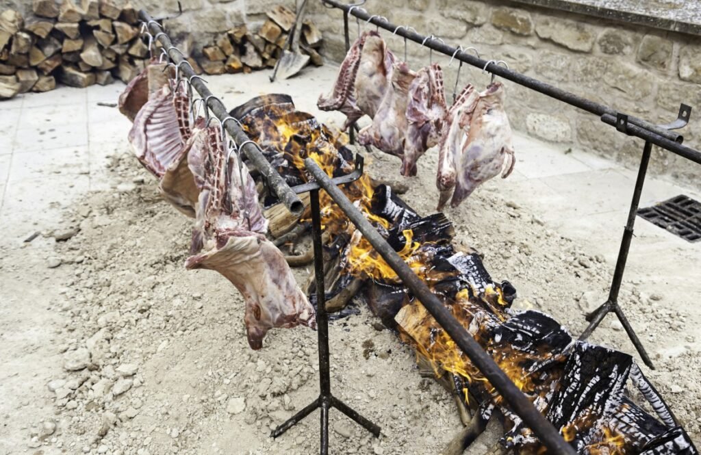 meat-hanging-over-a-roaring-fire-outdoors-2026-03-26-02-41-47-utc Raw meat cuts hung on metal racks above a wood fire outdoors, with a stone wall in the background.