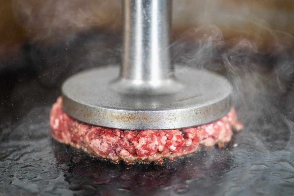 Ground beef patty being pressed on a hot grill by a metal burger press with steam rising nearby