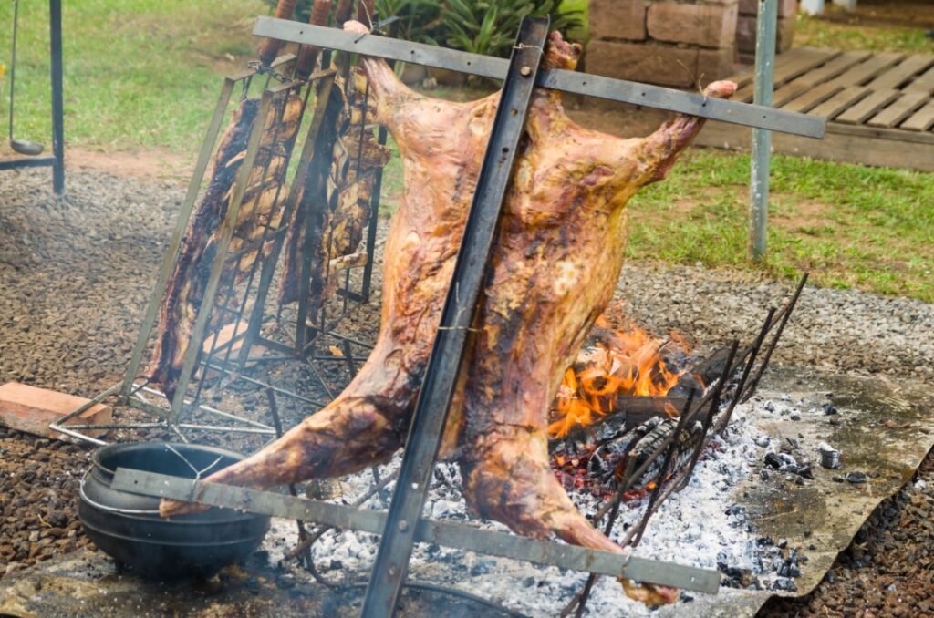 roasted-lamb-over-open-fire-in-rural-setting-2026-01-11-10-05-32-utc Two halves of a carcass being spit-roasted over a large outdoor fire, supported by metal frames in a backyard setting.