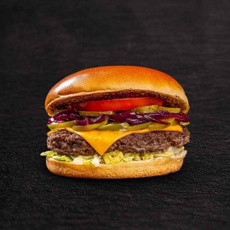 Juicy hamburger with beef patty, melted cheddar, pickle, tomato, red onion, and lettuce between a toasted bun on a dark background, viewed from the side.
