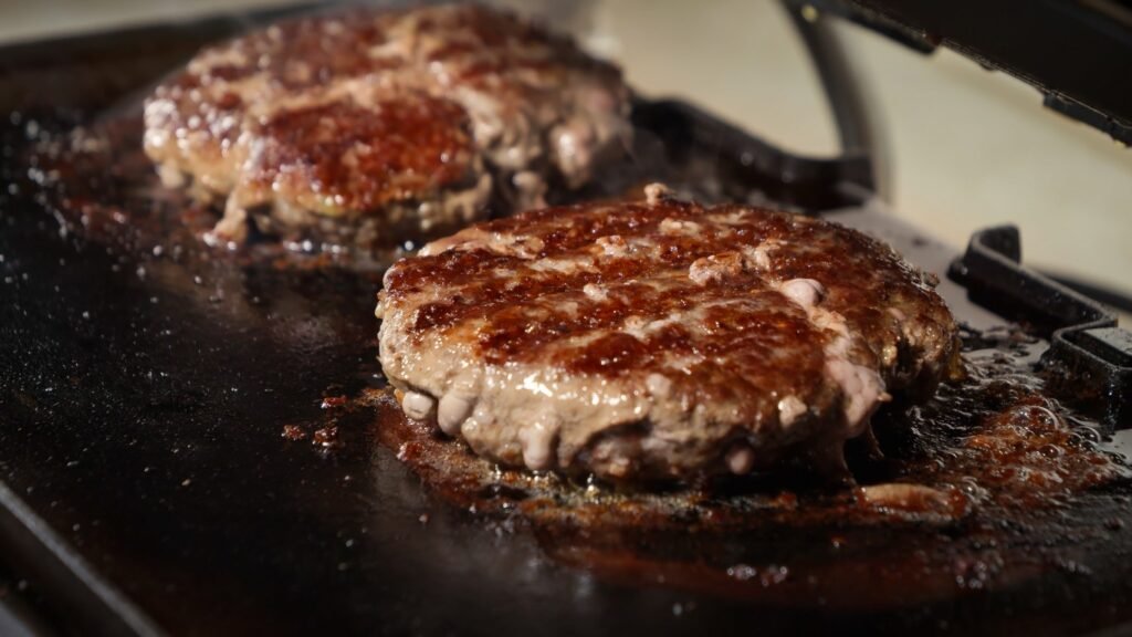 Two beef burger patties cook on a hot griddle, juices pooling around browned surfaces.