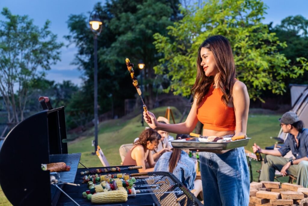 Woman in an orange top grills skewers at a backyard barbecue while friends relax on the lawn at dusk.