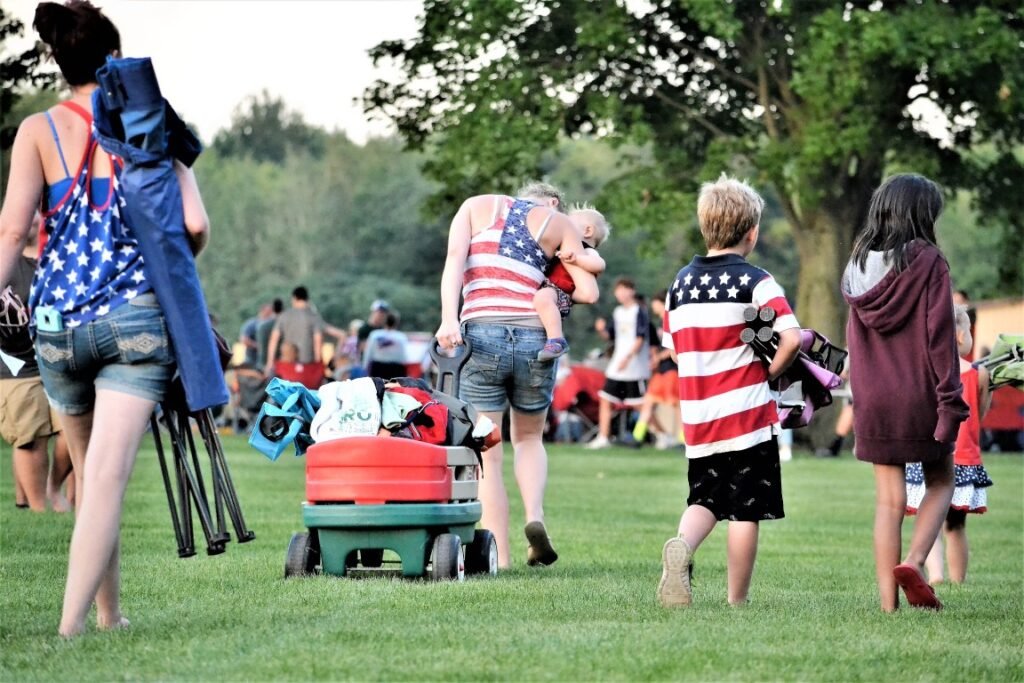 People at a park gathering on a sunny day; a woman carries a baby as others walk by with a wagon loaded with bags and gear, wearing patriotic outfits.
