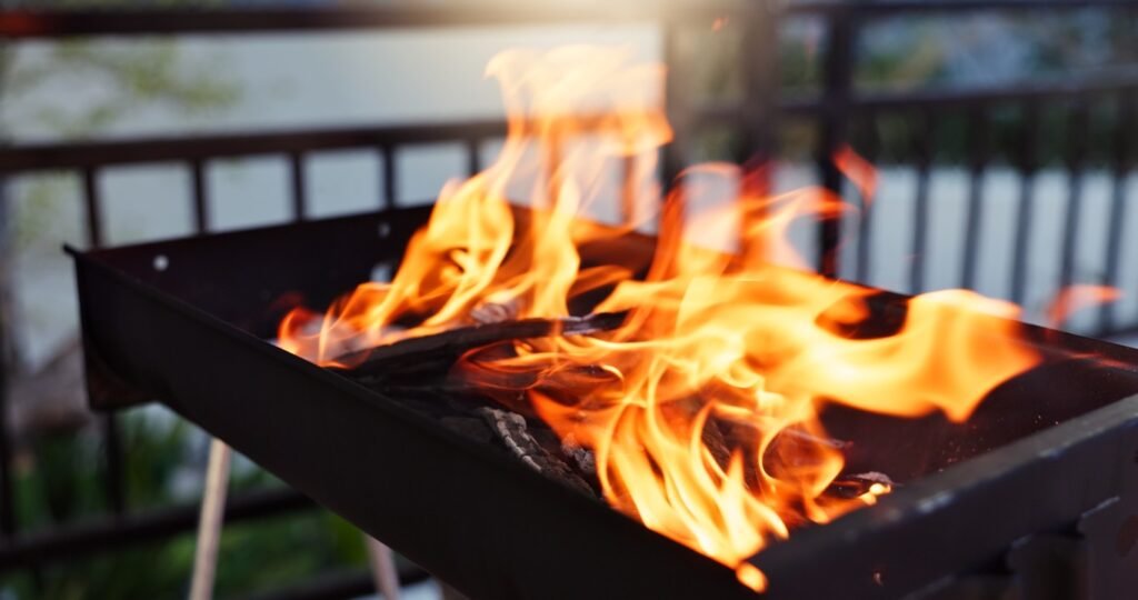 bbq-flame-and-grill-in-backyard-for-party-holida-2026-03-25-03-04-48-utc Flames lick a metal barbecue grill filled with burning charcoal on a deck.