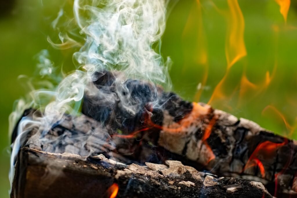 Burning firewood in the fire outdoor Log with glowing embers and curling white smoke rising from a campfire-like flame on charred wood