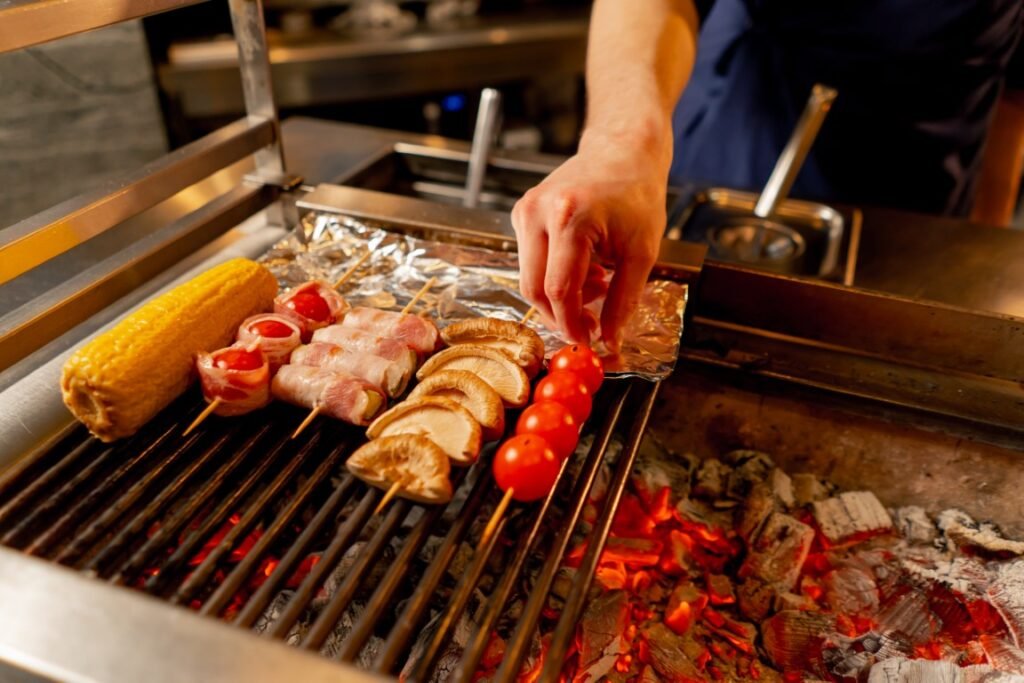 close-up in Japanese restaurant the chef grills various vegetables as a side dish for fish Hand placing bacon-wrapped skewers, mushrooms, and cherry tomatoes on a charcoal grill beside a corn cob drying nearby on foil-lined rack.