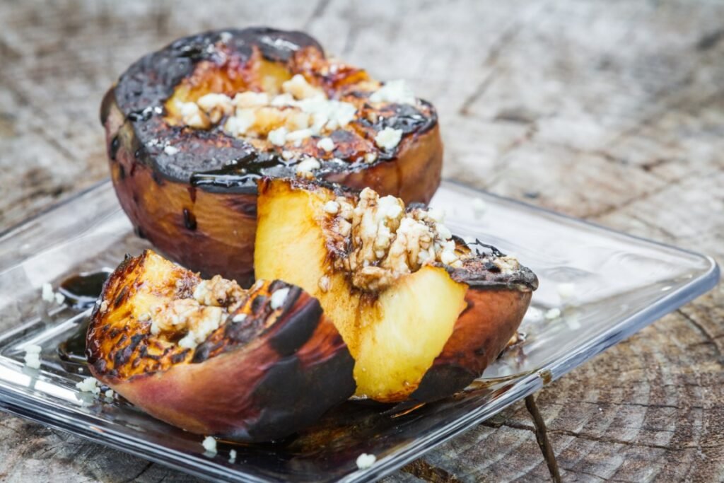 close-up-of-grilled-peaches-with-blue-cheese-and-a-2026-01-11-10-27-26-utc Grilled peach halves with chocolate glaze and crumb topping on a clear plate outdoors.
