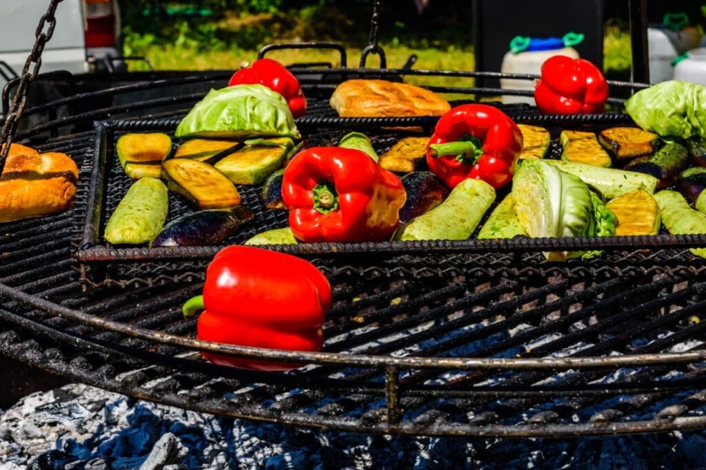 different-vegetables-cooking-on-grill-at-country-f-2026-03-26-01-23-32-utc Assorted vegetables, including red bell peppers, zucchini, cabbage, and eggplant, grilling on a barbecue over hot coals.