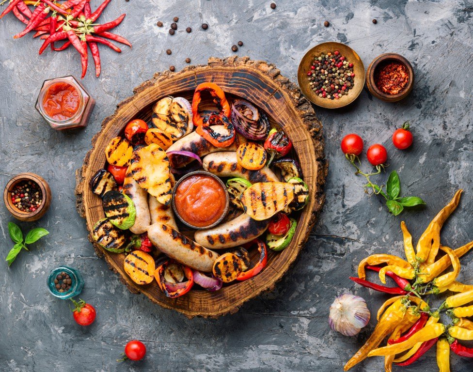 grilled-sausages-and-vegetables-on-wooden-cutting-2026-03-26-03-42-59-utc Assorted grilled sausages and vegetables on a rustic wooden platter with tomato dip, surrounded by chili peppers, garlic, and fresh herbs.