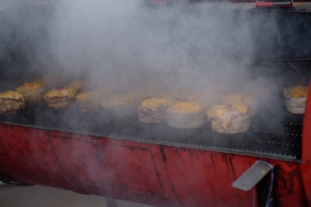 Handmade hamburger patties during the street food festival smoke while cooking Barbecue grill with several burgers cooking on a smoky grill, red grill body visible at the bottom.
