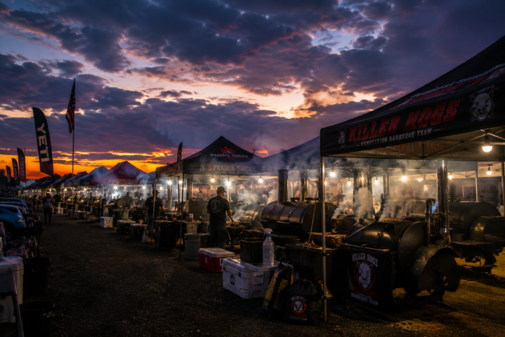 Sunset at a crowded outdoor BBQ competition with large smokers and tents set up along a row of stations.