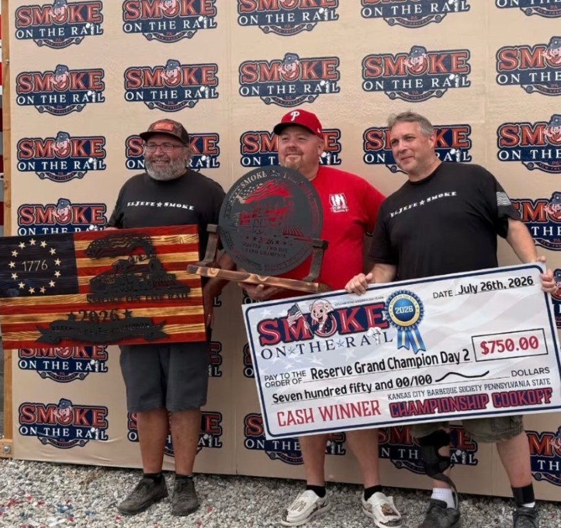 Three men pose with awards at Smoke on the Rail; left holds a wooden American flag plaque, center holds a circular metal trophy, right holds a large $750 prize check (Reserve Grand Champion Day 2).