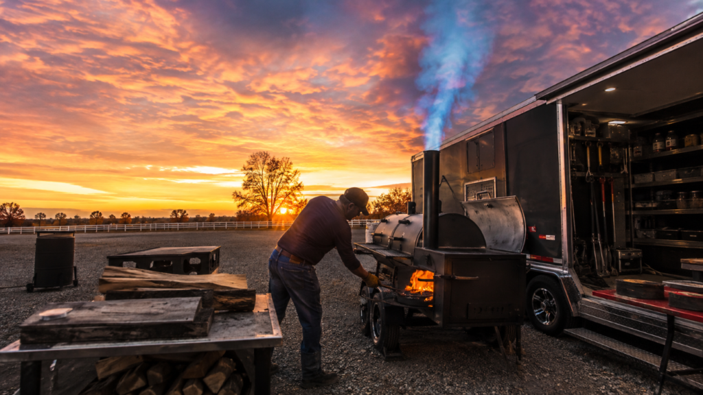 Man tending a wood-fired smoker attached to a trailer at sunset on a gravel lot, with flames visible in the firebox.