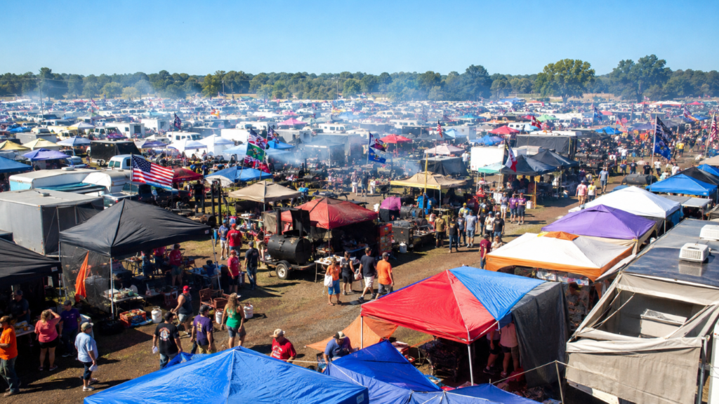 Large outdoor tailgate/festival scene with hundreds of colorful tents, stalls, and attendees under a clear blue sky.